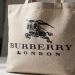 Close-up view of a textured beige linen tote bag featuring a black stenciled Burberry Equestrian Knight logo and "BURBERRY LONDON" text.